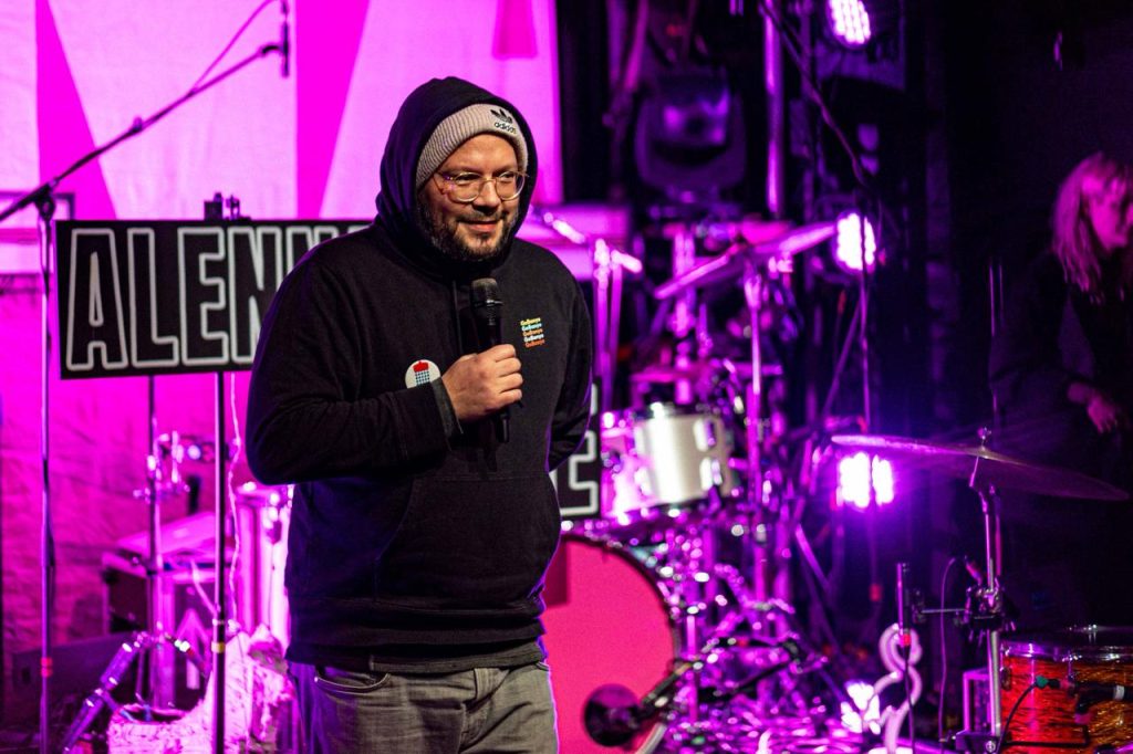 Auf einer Bühne mit Instrumenten im Hintergrund ist ein Mann in einem schwarzen Kapuzenpullover und einer Mütze zu sehen. Er hält ein Mikrofon in der Hand.