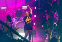 Schnee und Kälte: Diese Band ruft zur Unterstützung auf Eine Sängerin steht auf einer Konzertbühne. Sie singt in ein Mikrofon und hebt ihren Arm. Im Hintergrund sind weitere Bandmitglieder an der Gitarre und am Schlagzeug zu sehen.