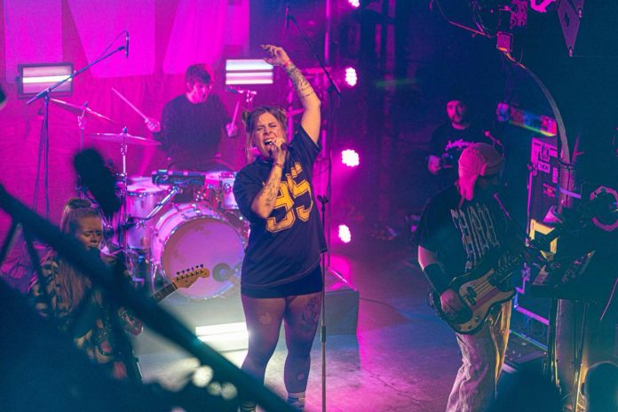 Eine Sängerin steht auf einer Konzertbühne. Sie singt in ein Mikrofon und hebt ihren Arm. Im Hintergrund sind weitere Bandmitglieder an der Gitarre und am Schlagzeug zu sehen.