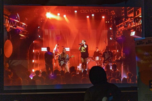 Eine Leinwand, auf der die &Uuml;bertragung eines Konzertes zu sehen ist.