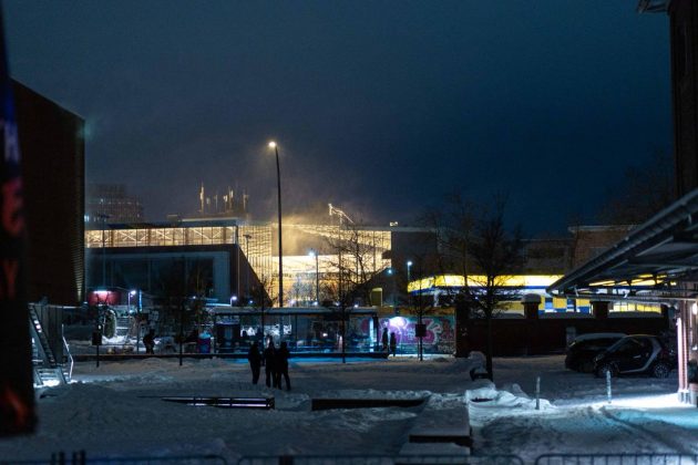 Ein schneebedeckter Platz bei Nacht. Lichter eines In den Lichtern aus den Fenstern eines Geb&auml;udes und einer Stra&szlig;enlaterne sieht man, dass es schneit.