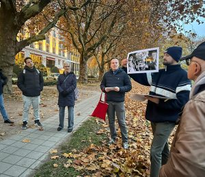 Mehrere Menschen stehen in einem Halbkreis in einem Park. Sie tragen Mäntel und Mützen. Ein Mann hält ein großes laminiertes Bild hoch. Die anderen Personen schauen auf das Bild in seiner Hand.
