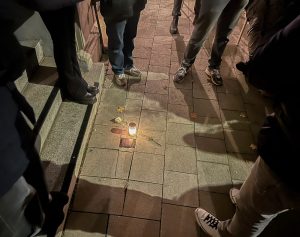 Mehrere Menschen stehen um einen Stoplerstein auf einem Bürgersteig herum. Es sind nur ihre Beine zu sehen. Neben dem Stolperstein steht eine Grabkerze. Daneben liegt eine weiße Rose.