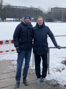 Zwei Männer stehen vor einem Fußballplatz. Es liegt Schnee im Hintergrund.