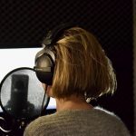 Wer spricht hier eigentlich? Hinterkopf einer Frau mit Kopfhörern vor ihr steht ein Mikrophon.