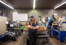 Von Studis gebaut: Ein Rennauto, das auch ohne Fahrer fährt Hawks Team Captain Charlotte Farwig lehnt an einem Tisch in der Werkstatt von Hawks Racing. Ihre Arme sind verschränkt, sie trägt eine Hawks Kappe. Links neben ihr steht der Rennwagen der Saison 2025. Rechts hinter ihr arbeitet ein Student an einer Werkbank.