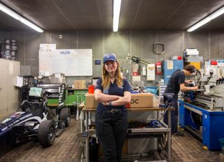 Von Studis gebaut: Ein Rennauto, das auch ohne Fahrer fährt Hawks Team Captain Charlotte Farwig lehnt an einem Tisch in der Werkstatt von Hawks Racing. Ihre Arme sind verschränkt, sie trägt eine Hawks Kappe. Links neben ihr steht der Rennwagen der Saison 2025. Rechts hinter ihr arbeitet ein Student an einer Werkbank.