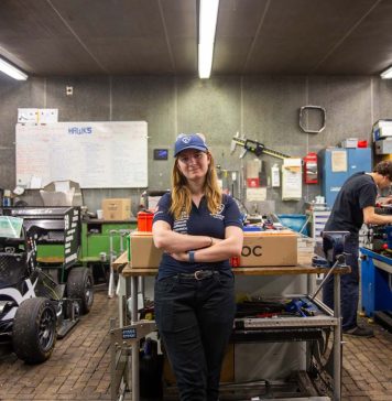 Von Studis gebaut: Ein Rennauto, das auch ohne Fahrer fährt Hawks Team Captain Charlotte Farwig lehnt an einem Tisch in der Werkstatt von Hawks Racing. Ihre Arme sind verschränkt, sie trägt eine Hawks Kappe. Links neben ihr steht der Rennwagen der Saison 2025. Rechts hinter ihr arbeitet ein Student an einer Werkbank.