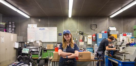 Von Studis gebaut: Ein Rennauto, das auch ohne Fahrer fährt Hawks Team Captain Charlotte Farwig lehnt an einem Tisch in der Werkstatt von Hawks Racing. Ihre Arme sind verschränkt, sie trägt eine Hawks Kappe. Links neben ihr steht der Rennwagen der Saison 2025. Rechts hinter ihr arbeitet ein Student an einer Werkbank.