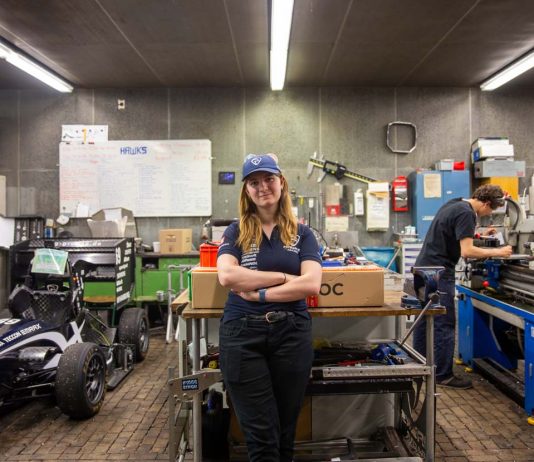 Von Studis gebaut: Ein Rennauto, das auch ohne Fahrer fährt Hawks Team Captain Charlotte Farwig lehnt an einem Tisch in der Werkstatt von Hawks Racing. Ihre Arme sind verschränkt, sie trägt eine Hawks Kappe. Links neben ihr steht der Rennwagen der Saison 2025. Rechts hinter ihr arbeitet ein Student an einer Werkbank.