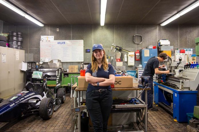 Hawks Team Captain Charlotte Farwig lehnt an einem Tisch in der Werkstatt von Hawks Racing. Ihre Arme sind verschränkt, sie trägt eine Hawks Kappe. Links neben ihr steht der Rennwagen der Saison 2025. Rechts hinter ihr arbeitet ein Student an einer Werkbank.