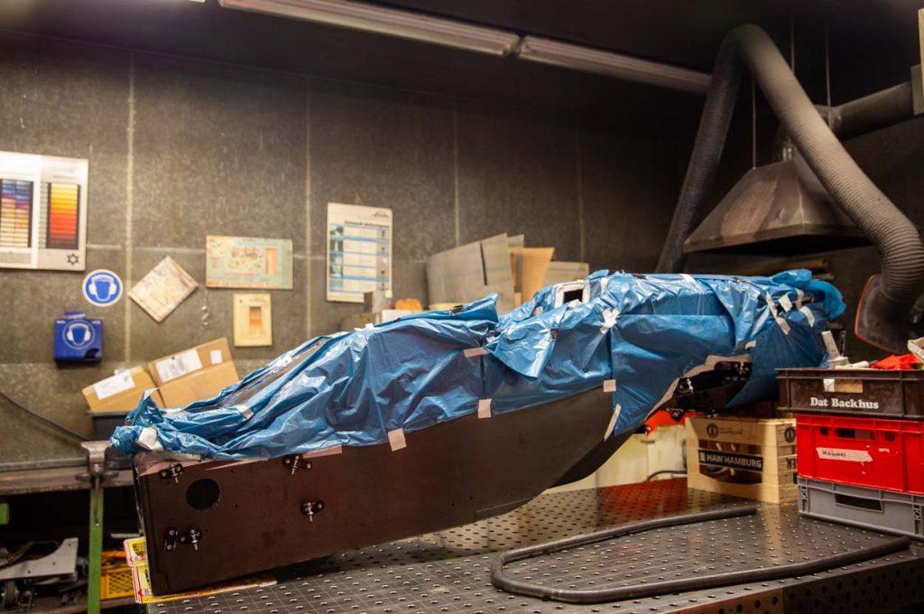 Das Gehäuse des Rennwagens liegt auf einem metallenen Tisch. Blaue Mülltüten kleben auf der oberen Hälfte des Autos. Dass Chassis ist länglich und soll auch autonom fahren können.