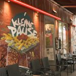 Kleine Pause, großer Trubel Die Außenfassade der Kleinen Pause. Auf der roten Wand ist ein Graffiti, dass eine Pommes Schale mit Ketchup zeigt. Darüber steht "Kleine Pause". Vor der Hauswand stehen Tische und Stühle unter einer Markise.