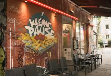 Kleine Pause, großer Trubel Die Außenfassade der Kleinen Pause. Auf der roten Wand ist ein Graffiti, dass eine Pommes Schale mit Ketchup zeigt. Darüber steht "Kleine Pause". Vor der Hauswand stehen Tische und Stühle unter einer Markise.