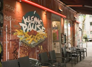 Kleine Pause, großer Trubel Die Außenfassade der Kleinen Pause. Auf der roten Wand ist ein Graffiti, dass eine Pommes Schale mit Ketchup zeigt. Darüber steht "Kleine Pause". Vor der Hauswand stehen Tische und Stühle unter einer Markise.
