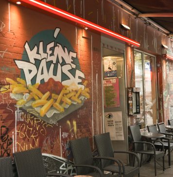 Kleine Pause, großer Trubel Die Außenfassade der Kleinen Pause. Auf der roten Wand ist ein Graffiti, dass eine Pommes Schale mit Ketchup zeigt. Darüber steht "Kleine Pause". Vor der Hauswand stehen Tische und Stühle unter einer Markise.