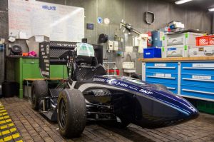 Ein dunkelblauer Rennwagen steht in der Werkstatt der Hawks. Das Gehäuse glänzt, die Reifen sind abgenutzt und dreckig. Hinter dem Auto hängt ein Whiteboard an der Wand mit Daten und Informationen zu Hawks Racing.