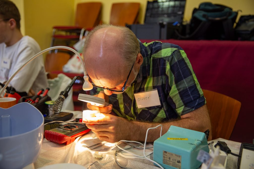 Das Bild zeigt älteren Mann, der im Licht einer gebogenen Schreibtischlampe und mithilfe einer Lupe ein weißes Gehäuse betrachtet. Er sitzt vorgebeugt an einem Tisch, auf dem viel Werkzeug liegt.