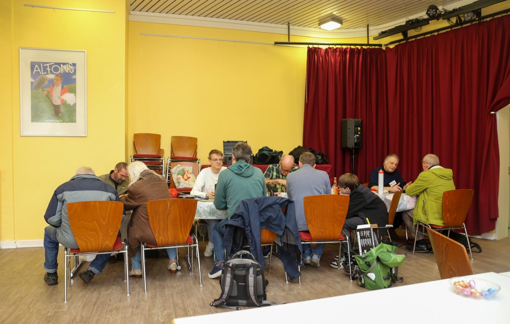 Das Bild zeigt verschiedene Personen, die sich an einer Reihe von Tischen gegenübersitzen. Es werden mehrere Reparaturen gleichzeitig durchgeführt, die Personen im Bild unterhalten sich. Sechs der insgesamt zehn abgebildeten Personen sind nur mit dem Rücken zu sehen.