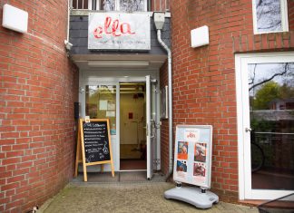 Wo (fast) alles ein zweites Leben bekommt Das Foto zeigt den Eingang zum Café ella. Die Tür steht offen. Das Gebäude ist ein Backsteinhaus. Neben der geöffneten Tür stehen Aufsteller, die Flyer und einen Teil der Speisekarte zeigen.