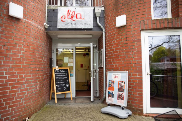 IMG_8706 Das Foto zeigt den Eingang zum Café ella. Die Tür steht offen. Das Gebäude ist ein Backsteinhaus. Neben der geöffneten Tür stehen Aufsteller, die Flyer und einen Teil der Speisekarte zeigen.