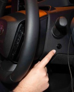 Ein schwarzes Lenkrad mit einem Panikknopf daneben. Der Taxifahrer hält seinen ausgestreckten Finger neben den Knopf.