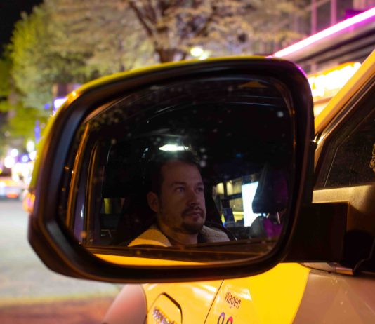 „Beichtvater und Therapeut”: Unterwegs mit einem Taxifahrer Die Spiegelung von dem Taxifahrer Daniel Lebastchi in dem Seitenspiegel seines gelben Taxis. Im Hintergrund ist die Reeperbahn zu erkennen.