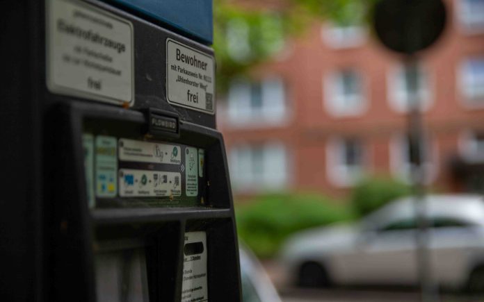 Nahaufnahme eines Parkautomaten bei Tag. Im Hintergrund verschwommen: ein Straßenschild und graue Autos.