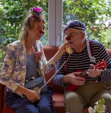 Ein Clown im Pflegeheim: „Mögt ihr Plattdeutsch?“ „Jaaaaa!“ Martin Schneider-Schall und Franziska Heuschkel sind Clowns im Pflegeheim. Sie tragen bunte Klamotten sowie eine rote Plastiknasen und sitzen auf einem Sofa zusammen.