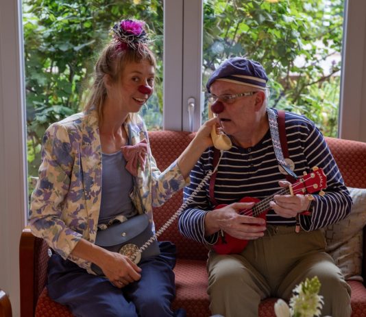 Ein Clown im Pflegeheim: „Mögt ihr Plattdeutsch?“ „Jaaaaa!“ Martin Schneider-Schall und Franziska Heuschkel sind Clowns im Pflegeheim. Sie tragen bunte Klamotten sowie eine rote Plastiknasen und sitzen auf einem Sofa zusammen.