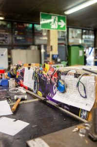 Mehrere Schutzbrillen und Kopfhörer hängen über einem Regalbrett, auf dem ein Bild von einem Rennwagen klebt. Mehrere Zettel sind an das Brett geklemmt und davor liegt ein Hammer auf einer schwarzen Unterlage. Im Hintergrund befindet sich ein großes Exit-Schild.