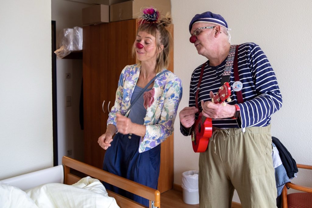Zwei Clowns singen ein Lied am Bett eines Senioren im Pflegeheim. Ein Clown spielt auf einer roten Ukulele.