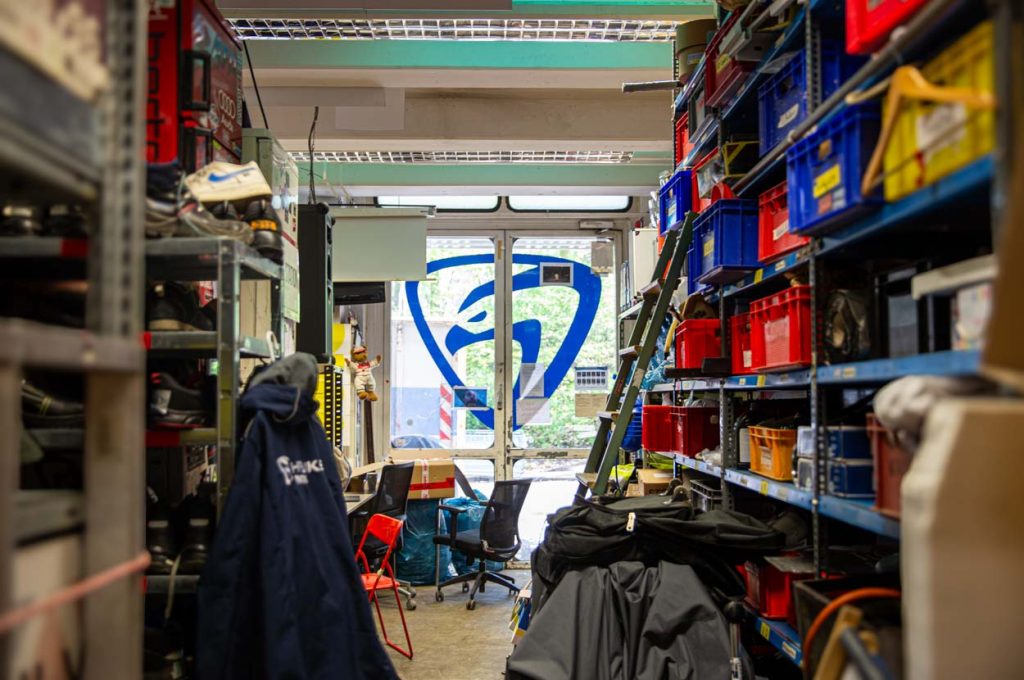 Auf einer gläsernen Doppeltür ist das Logo der Hawks groß gedruckt. Links und rechts stehen volle Regale, in denen Plastikkisten stecken. An dem rechten regal lehnt eine metallene Leiter. Im linken Regal stehen Schuhe und eine Jacke mit Hawks Emblem ist daran angehängt.