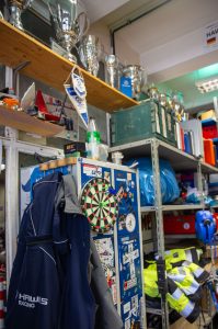 An einem kleinen blauen Schrank kleben mehrere Sticker, im Hintergrund liegen neongelbe Warnjacken. Am Schrank hängt eine dunkle Hawks Racing Jacke. Hoch über dem Schrank ist ein hölzernes Regalbrett, auf dem silberne, glänzende Pokale stehen. 