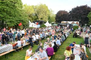 Viele Menschen sitzen an mehreren langen Tischen auf einer Wiese und essen.