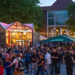 Kostenlos Kultur! Die Altonale startet Das Bild zeigt viele Menschen, die in der Abenddämmerung draußen stehen und tanzen. Im Hintergrund sind bunt beleuchtete Gebäude zu sehen.