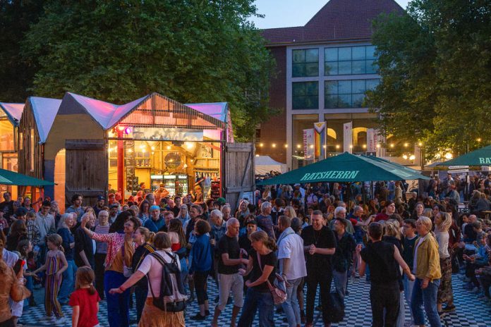 Das Bild zeigt viele Menschen, die in der Abenddämmerung draußen stehen und tanzen. Im Hintergrund sind bunt beleuchtete Gebäude zu sehen.