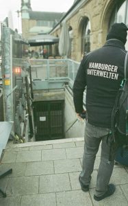 Ein Mann steht mit dem Rücken zum Betrachter am Fuße eines Abgang in einen Bunker. Auf seiner Jacke steht: "Hamburger Unterwelten".