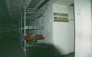 Zweier Sitzbänke in einem dunklen Bunker neben einer Wand mit der Aufschrifft: "Zum 2.+3. Geschoss und Ausgang"