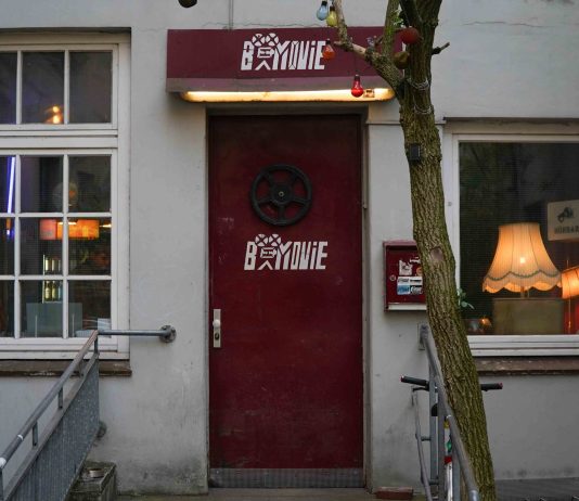Kino, günstiger als Netflix Eine Neonröhre wirft von oben Licht auf eine rote Tür, in deren Mitte eine Filmrolle hängt und die Aufschrift "B-Movie" zu lesen ist.