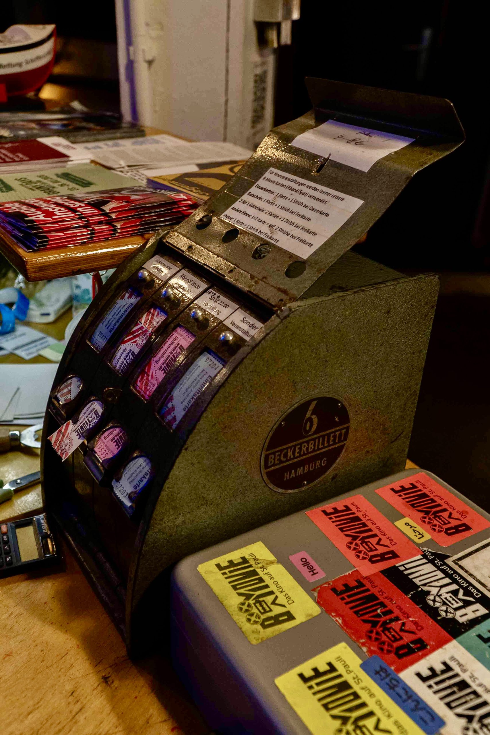 Eine graue, eiserne Kasse mit bunten Kinotickets aus Papier steht auf einem Tresen. Rechts daneben eine Geldkassette, die mit Stickern beklebt ist.