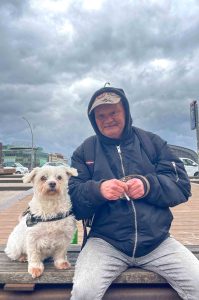 Chris sitzt mit seinem Hund Struppi auf einer Bank. Zwischen seinen Fingern hält er eine Zigarette.