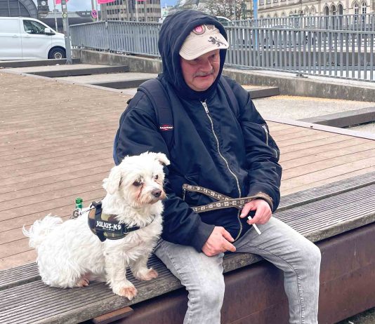 Früher obdachlos, heute Stadtführer Chris sitzt mit seinem Hund Struppi auf einer Bank. Zwischen seinen Fingern hält er eine Zigarette.