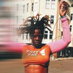 „Deutschland ist das härteste Casting der Welt“ Schwarze Frau mit abstehenden Locks und orange-pinken Crop-Top im Vordergrund. Hände sind hochgehoben und ein verschwommener Effekt zieht die Frau in die Länge. Sie steht vor einem weißen Gebäude.