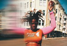 „Deutschland ist das härteste Casting der Welt“ Schwarze Frau mit abstehenden Locks und orange-pinken Crop-Top im Vordergrund. Hände sind hochgehoben und ein verschwommener Effekt zieht die Frau in die Länge. Sie steht vor einem weißen Gebäude.