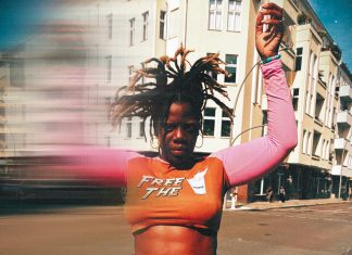 „Deutschland ist das härteste Casting der Welt“ Schwarze Frau mit abstehenden Locks und orange-pinken Crop-Top im Vordergrund. Hände sind hochgehoben und ein verschwommener Effekt zieht die Frau in die Länge. Sie steht vor einem weißen Gebäude.
