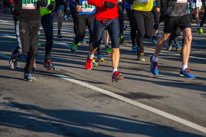 Mehrere Menschen laufen bei einem Marathon über eine Asphalt-Straße. Das Bild ist oben abgeschnitten, sodass man nur die untere Häkfte der Menschen sieht. Sie tragen Sportkleidung und haben teilweise Schilder mit Zahlen auf ihren T-Shirts.
