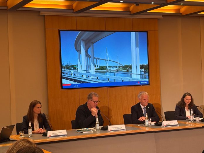 IMG-20260610-WA0023 (1) 4 Personen sitzen in einer Pressekonferenz. auf einem Bildschirm hinter ihnen ist eine Visualisierung des Entwurfs der neuen Köhlbrandbrücke zu sehen.