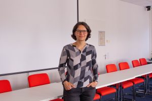 Das Bild zeigt Anna Franze, Erziehungswissenschaftlerin, in einem Raum. Sie steht vor einem Tisch mit roten Stühlen.