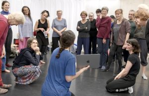 In einem Tanzsaal stehen Menschen in einem Kreis. Auf dem Boden sitzt die Choreografin mit einem Laptop in der Hand.